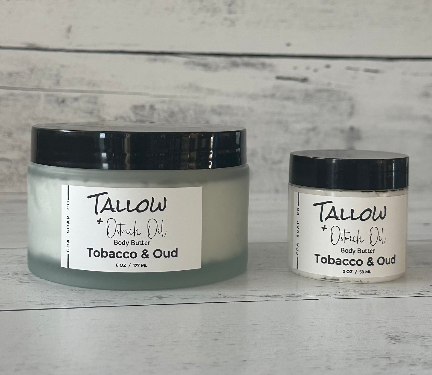 Two jars of Tallow + Ostrich Oil body butter on a wooden surface with a gray background.