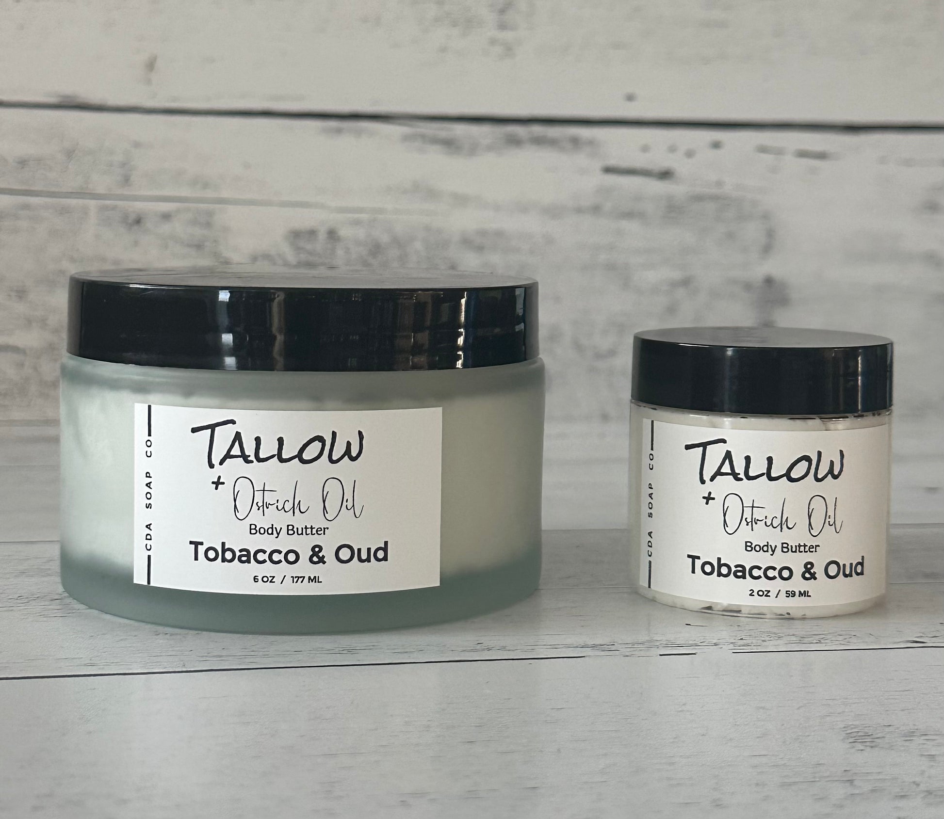 Two jars of Tallow + Ostrich Oil body butter on a wooden surface with a gray background.