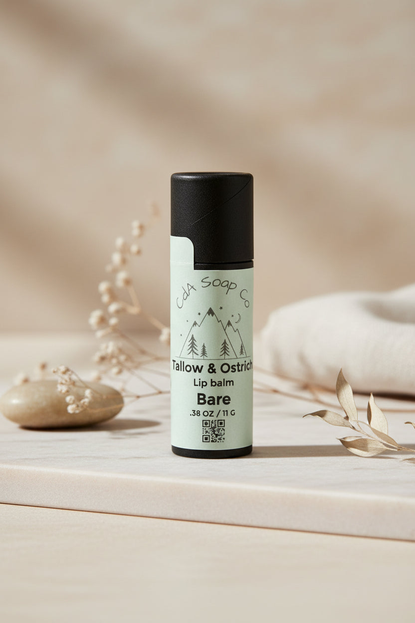 Black Kraft board lip balm tube with white label for an unscented tallow and ostrich oil lip balm. Light back ground with small subtle accents of baby’s breath, a stone and part of a blanket 