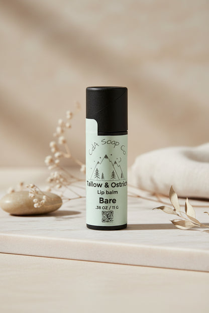 Black Kraft board lip balm tube with white label for an unscented tallow and ostrich oil lip balm. Light back ground with small subtle accents of baby’s breath, a stone and part of a blanket 