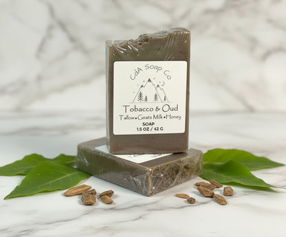 Bar of soap with packaging labeled 'Tobacco & Oud' on a wooden surface with leaves and seeds.
