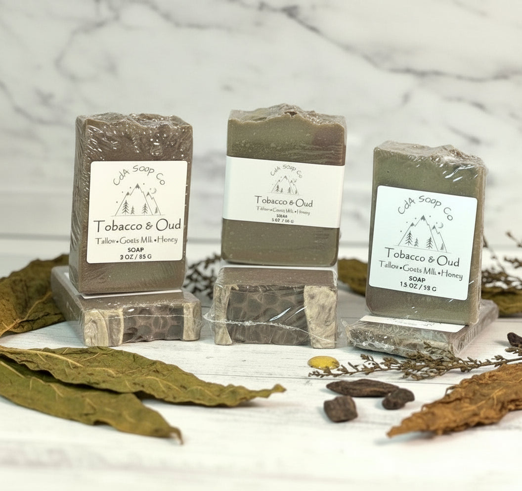 Three bars of Tobacco & Oud soap on a wooden surface with dried leaves and herbs.