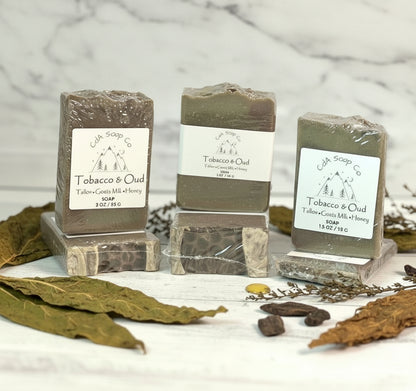 Three bars of Tobacco & Oud soap on a wooden surface with dried leaves and herbs.