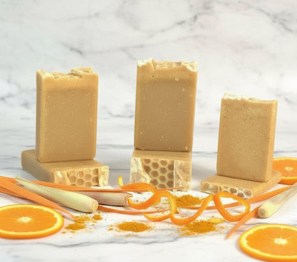 Three bars of brown soap on stands with orange slices and a honeycomb on a light marble background