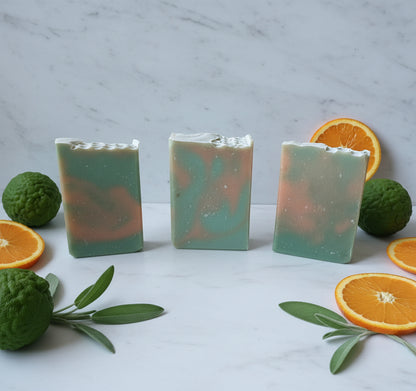 Three bars of soap with green and orange hues on a marble surface with oranges and limes.