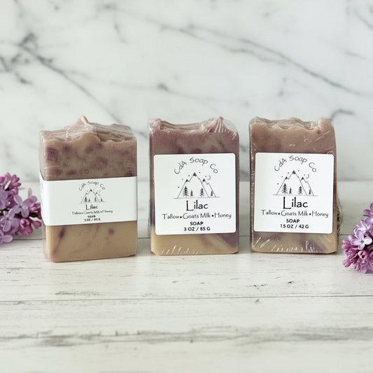 Three bars of lilac soap from CA Soap Co on a wooden surface with purple flowers.