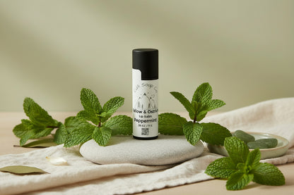 Black Kraft board peppermint lip balm tube. Light back ground with peppermint leaves, a stone and a blanket 