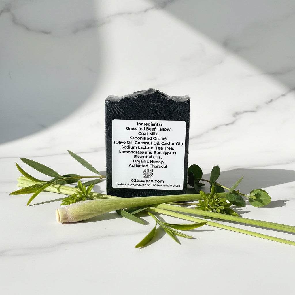Black soap bar with ingredients label on a white marble surface with green leaves.