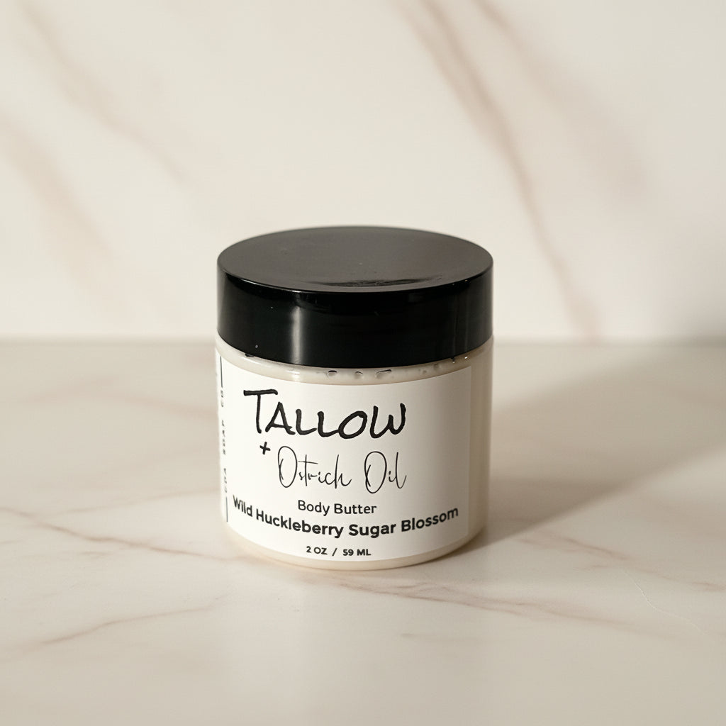 A small jar with a black lid labeled 'TALLOW + Ostrich Body Butter Wild Huckleberry Sugar Blossom'. The jar is placed against a wooden background.