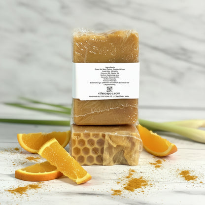 Two bars of soap with a honeycomb pattern, surrounded by orange slices and lemongrass on a light marble surface.