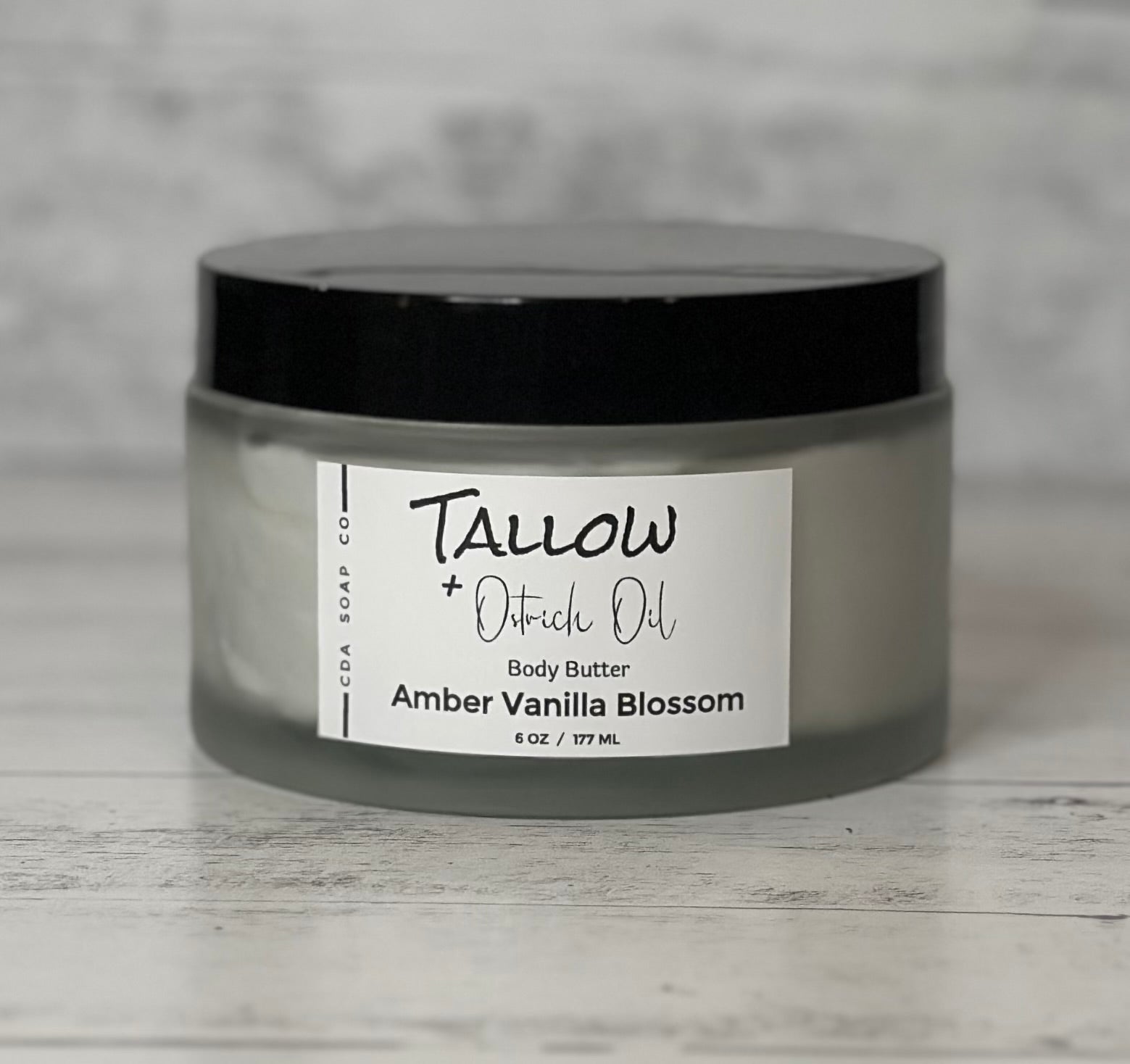 A jar of Tallow + Ostrich Body Butter with a label that includes the product name and a brief description. The jar is made of frosted glass with a black lid, placed on a wooden surface.