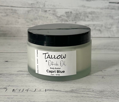 An image featuring a jar of Capri Blue Tallow & Ostrich Body Butter. The larger 6 oz jar is in a frosted white glass jar with a black lid, against a light wood background 