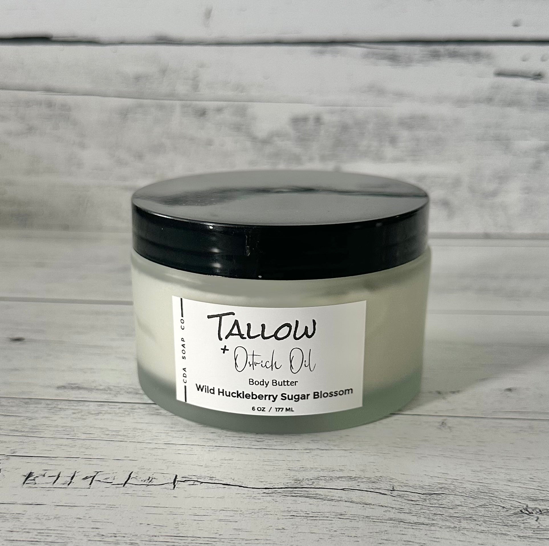 A frosted white glass jar with a black lid labeled 'TALLOW + Ostrich Body Butter Wild Huckleberry Sugar Blossom'. The jar is placed against a wooden background.