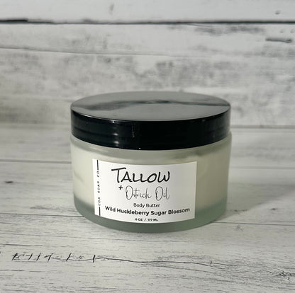 A frosted white glass jar with a black lid labeled 'TALLOW + Ostrich Body Butter Wild Huckleberry Sugar Blossom'. The jar is placed against a wooden background.