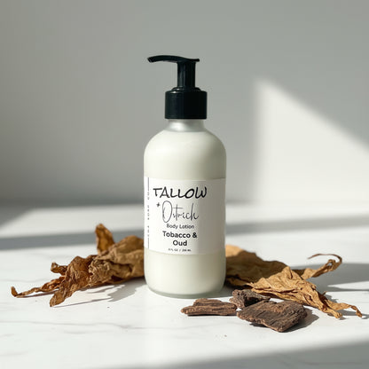 Body lotion bottle labeled 'Tobacco & Oud' on a light surface with dried leaves and tobacco.